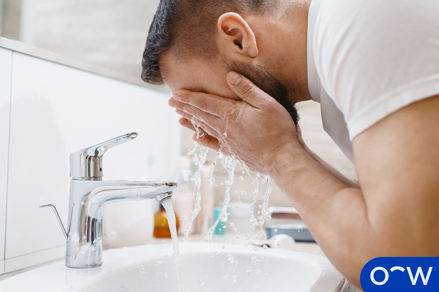 A person splashing water on their face over a bathroom sink using water from a running tap to clean their eyes.